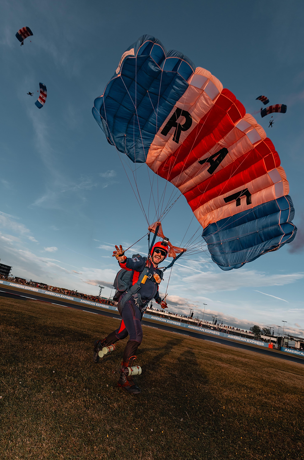 An RAF Falcons parachute display team member in a navy blue flight suit and red helmet lands on a Silverstone racetrack showground, gesturing with a peace sign under a large red, white, and blue parachute bearing the letters 'RAF' on the white panels. Four more RAF Falcons circle above, ready to land.
